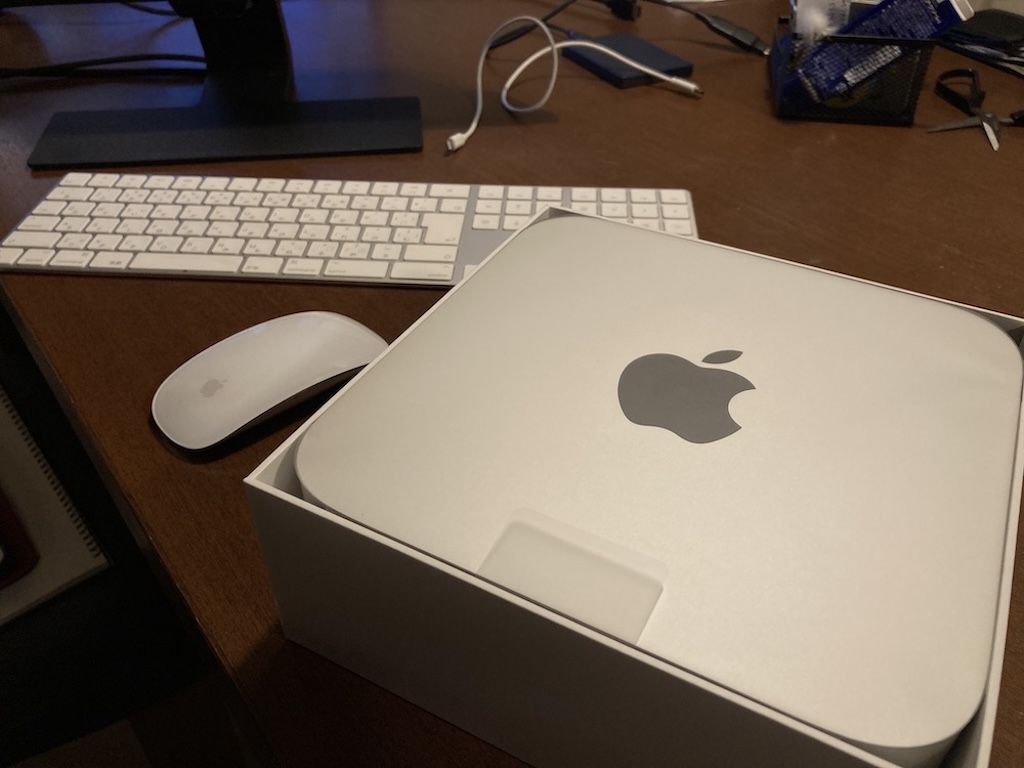 木製のデスク上に置かれた、白い外箱に入ったApple Mac mini(シルバー、トップにAppleロゴ)。その左隣にはMagic MouseとMagic Keyboardが並んでおり、背景にはモニターのスタンドやケーブル類が見える、開封直後の様子。