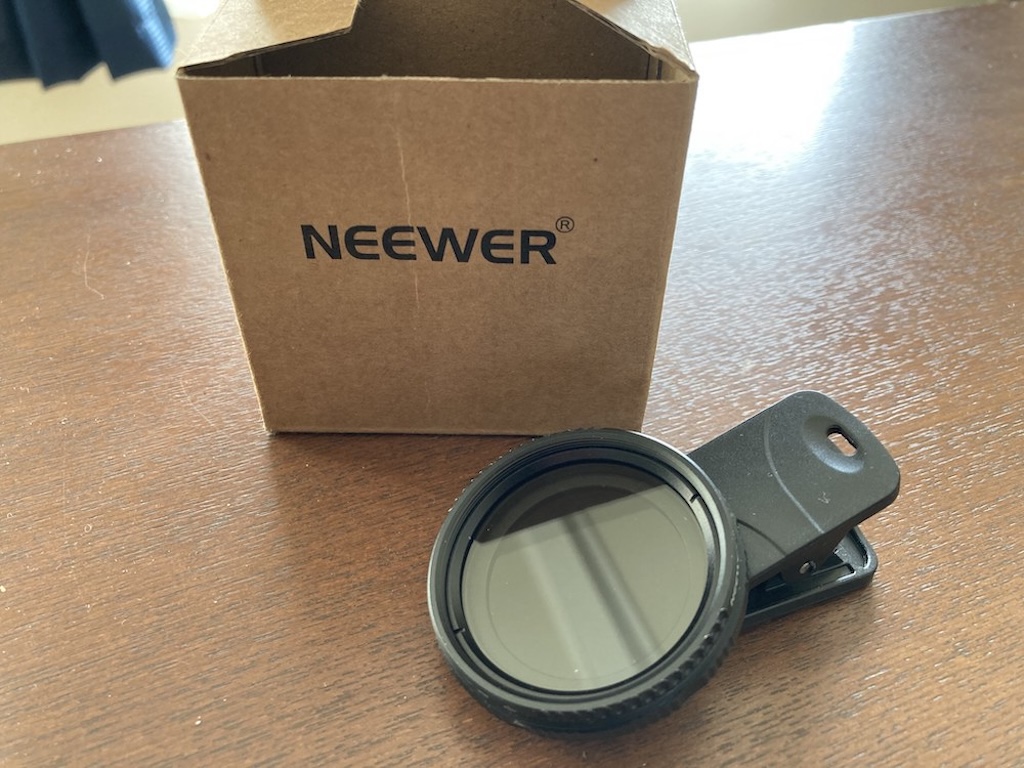 木目調のテーブルの上に置かれた、黒いクリップオン式のスマートフォン用NDフィルターと、その奥にある「NEEWER」のロゴが印刷されたクラフト紙の化粧箱。